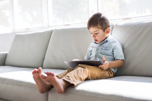 Menino com tablet digital sentado no sofá, em casa fundo interior — Fotografia de Stock
