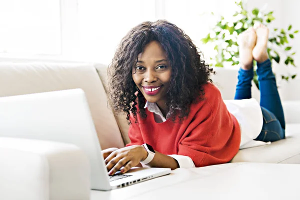 Jolie jeune femme noire utilisant un ordinateur portable tout en se relaxant sur un canapé à la maison — Photo