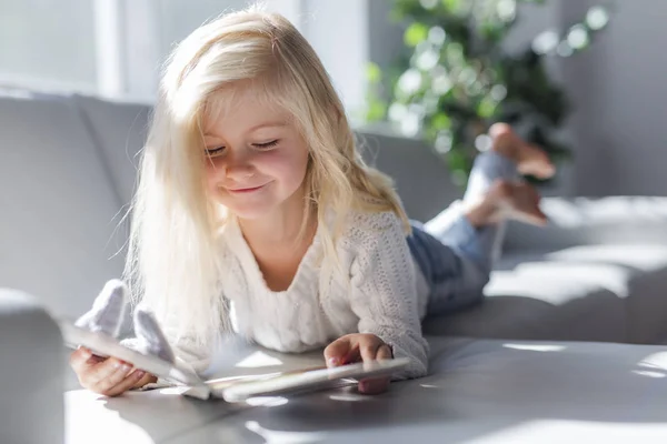 Очаровательная маленькая девочка читает книгу на белом диване — стоковое фото
