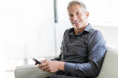 Portrait of mature man relaxing at home in sofa and cellphone clipart