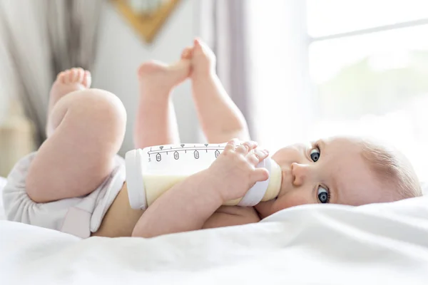 Красивая малышка пьет воду из бутылки, лежащей на кровати. Детский подгузник в детской комнате . — стоковое фото