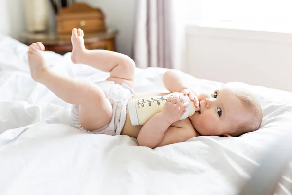 Jolie petite fille boit de l'eau du biberon couché sur le lit. Couche portée enfant dans la chambre d'enfant . — Photo