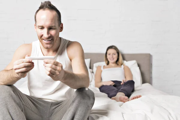 Homem sentado na cama com mulher grávida por trás olhar para o teste de gravidez — Fotografia de Stock
