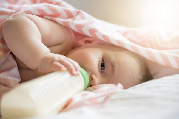 Маленька дівчинка лежить на білому ліжку і п'є молоко з пляшки — стокове фото