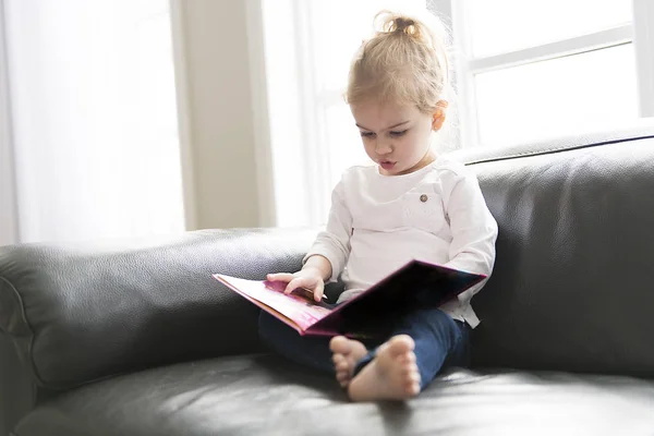 Портрет милої маленької дівчинки сидить на дивані у вітальні насолоджуйтесь читанням великої книги — стокове фото