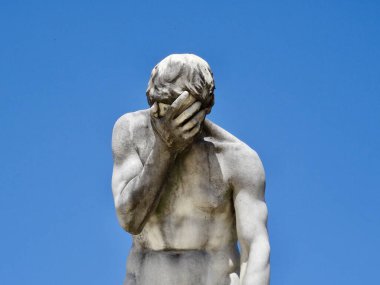 Eski taş heykel erkek figürü, yüzü sanki bir facepalm yapıyormuş gibi tutuyor.              