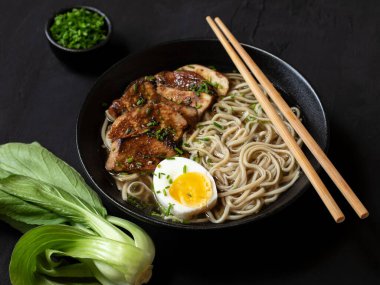 Siyah arka planda Ramen Erişte ve Tavuk Çorbası. Izgara tavuk, erişte ve siyah bir kasede yumurta. Taze soğan ve bok choy bir yana. Kapat..