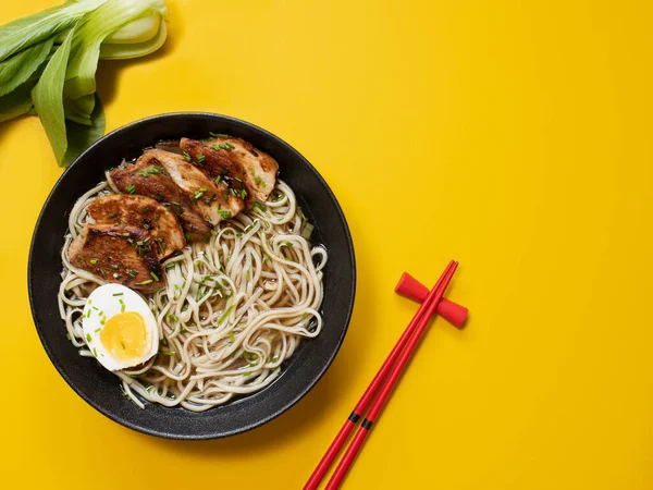 Sarı arka planda Ramen Erişte ve Tavuk Çorbası. Izgara tavuk, erişte ve siyah bir kasede yumurta çubuklar ve bok çuvalı. Üst Manzara. Kapat..