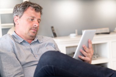 Yetişkin bir adam kanepede oturuyor ve evdeki tabletten haber okuyor.