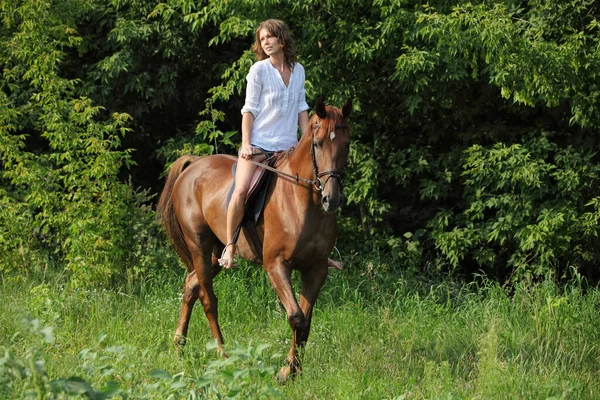 Beautiful blond girl riding sports dressage horse in summer forest