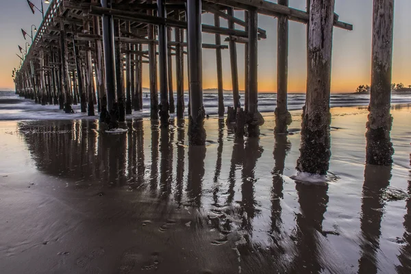 干潮から濡れた砂として海の地平線の後ろに最後の光による退色が日没時木造構造を反映します — ストック写真