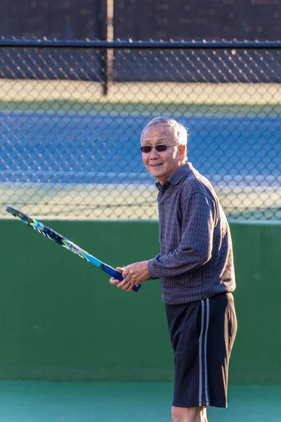 Smiling Chinese elderly man having fun while enjoying a game of tennis with racquet in hand.