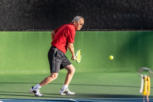 Focused Chinese elderly man keeping eyes on the ball as he prepares to hit a backhand  during a game of tennis.