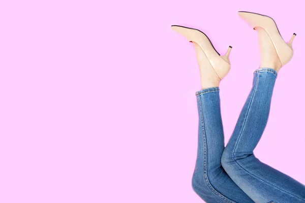 Beautiful Woman Feet & Slim Legs in Beige Medium High Heels on Pastel Pink. Portrait of Sexy Legs. Young Female Wearing Jeans (Blue Pants) in Stylish Shoes Raised Up Isolated on Pink Color Background.