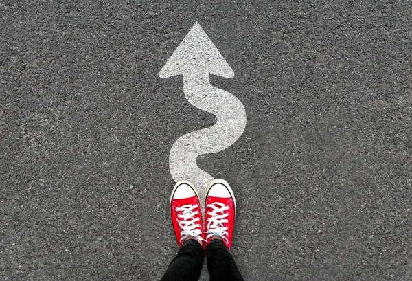 Feet and white arrow sign go straight on road background. Top view of woman. Forward movement and motivation idea concept. Selfie of foot and legs in red sneaker shoes on pavement floor from above.