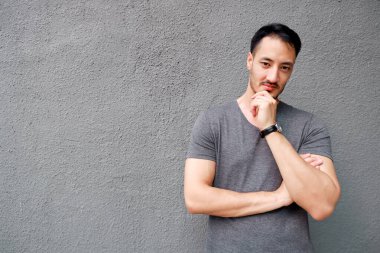 Gri arka planda duran mutlu genç adamın portresi. Tişört giymiş yakışıklı bir erkek model. Gri elleri çapraz, seksi kolları ve düşünme fikri. Moda genç adam düşünüyor ve kameraya bakıyor..