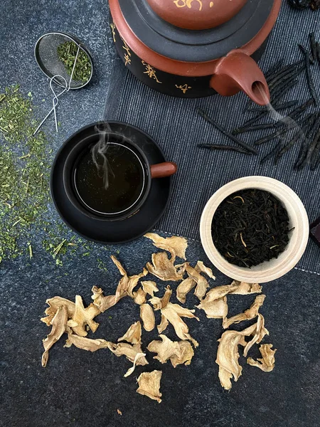 Ingredients for a healthy cold tea. Dried ginger and mint. Black tea and Chinese kudin tea. Chinese teapot and a small cup on a dark stone textured surface with a top view.