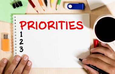 Hesap makinesi ve ofis malzemelerinin yanında beyaz bir arkaplan üzerinde PRIORITIES yazılı not. İş konsepti.