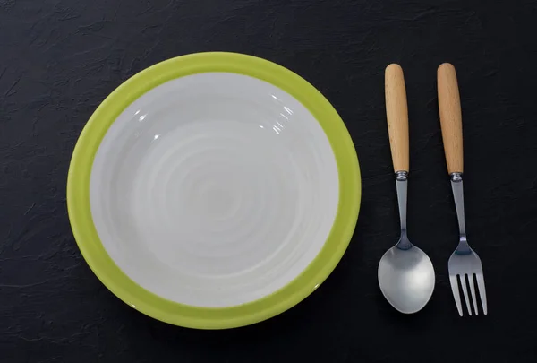用勺子和叉子固定在旧的黑色木制台面上的空白的白色瓷盘 — 图库照片