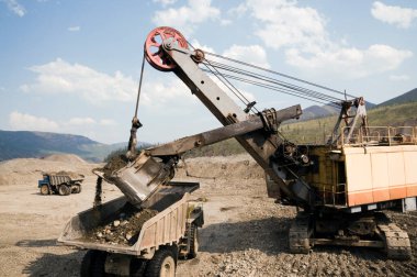 Güneşli bir yaz gününde dağlık bir ormanda kazıcı kazıcı ve maden kamyonları kullanarak bir çöp kamyonunun cesedine dağ toprağı yüklemek. Altın madenciliği endüstrisi.