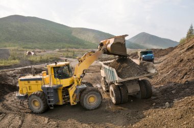 Dağlık bir ormandaki tekerlek yükleyicisi bir maden kamyonunu kovayla doldurup, dağ toprağını içine boşaltıyor. Altın madenciliği endüstrisi.