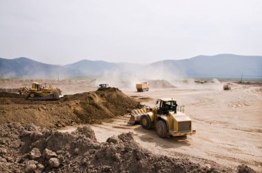 Güneşli bir yaz gününde açık bir alanda toprak işleri. Kargo kamyonları, kazıcılar, tekerlek yükleyiciler ve buldozerler aynı anda çalışır..