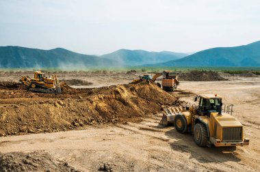 Güneşli bir yaz gününde açık bir alanda toprak işleri. Kargo kamyonları, kazıcılar, tekerlek yükleyiciler ve buldozerler aynı anda çalışır..