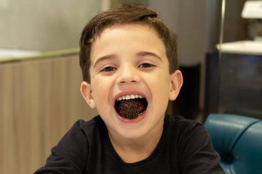 Child with sweet brigadeiro (brigadier) in mouth in a Brazilian bakery. clipart
