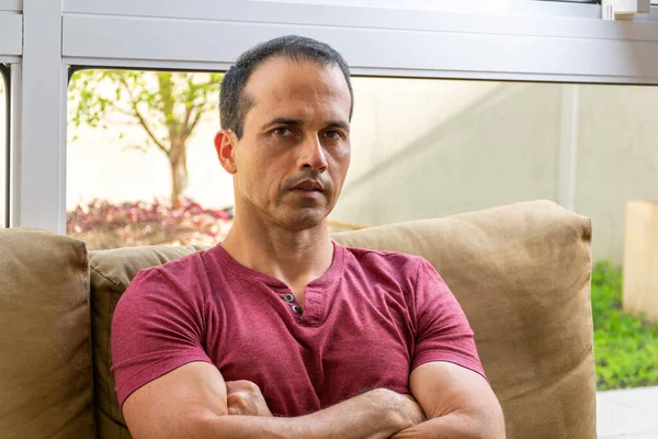 Kollarını kavuşturup kameraya bakan orta yaşlı bir adamın kanepede oturması..