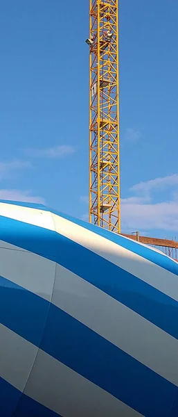 Close-up of the back of a blue white striped cement mixer truck in front of a yellow crane and the blue sky