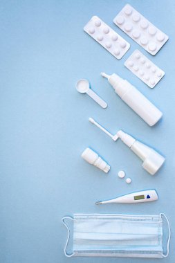 Tıbbi maske, beyaz termometre, haplar, mavi arka planda burun spreyi ve boğaz spreyi olan şişeler.