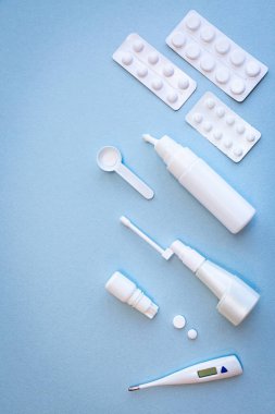 Beyaz termometre, haplar, burun spreyi şişeleri ve mavi arka planda boğaz spreyi.