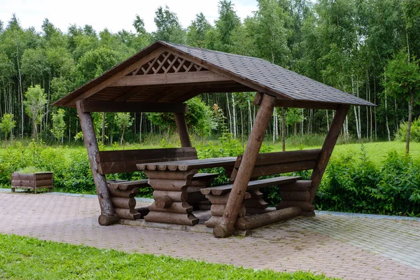 A roadside gazebo for tourists, made of logs, stands on a wide cobblestone path against the backdrop of a lawn with green grass and dense forest.