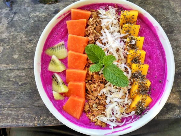 Taze papaya, mango, kivi, parçalanmış hindistan cevizi, chia tohumu, granola ve ahşap masa arkaplanlı bir nane filizi ile kaplı mor ejderha meyve suyu kasesi.