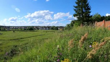 Beyaz bulutlar geçiyor. Çayırda yeşil çimenler dalgalanır ve nehir sola doğru akar. Mavi gökyüzü olan açık bir gün. Arka planda köy evleri var. Zaman Uygulaması.
