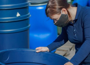 Koruyucu maskeli bir kadın mavi plastik bir fıçıyı değerlendiriyor..