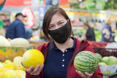 Çarşıda tıbbi maske takan bir kadın elinde bir karpuz ve kavun tutuyor.