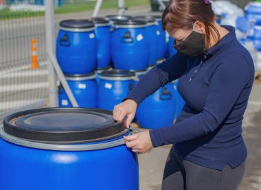 Siyah tıbbi maskeli bir kadın plastik bir fıçı açıyor..
