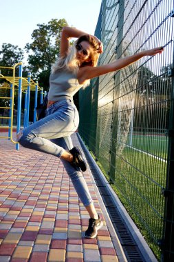 Mutlu genç bayan dışarıda eğleniyor, atlıyor ve tek başına dans ediyor. Farklı ekipmanlara sahip spor stadyumu. Uzun sarı saçlı, yüzünde güneş gözlüğü olan. Kot pantolon, spor ayakkabı ve gri bluz giyiyor..