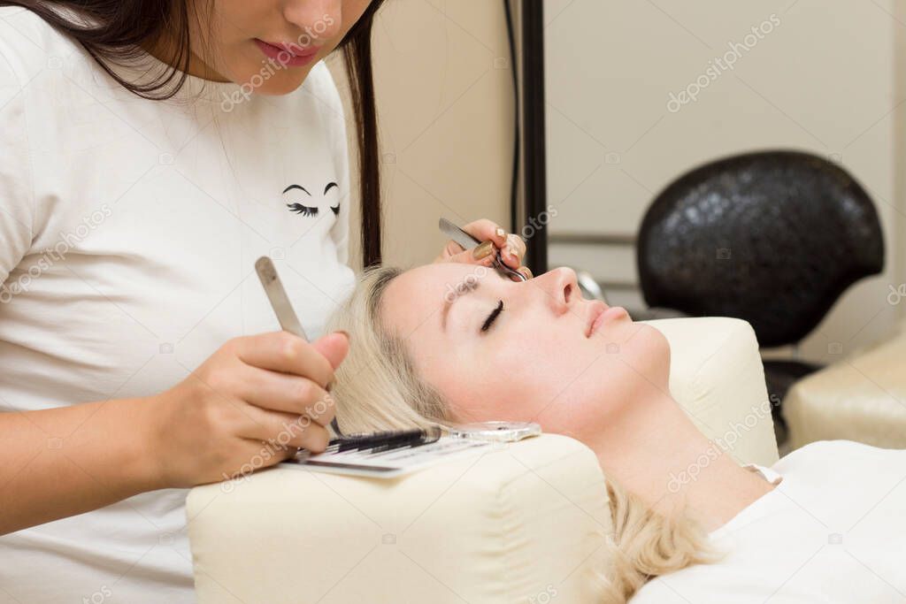 the master performs the procedure of eyelash extension with tweezers. A young woman is lying on a couch with her eyes closed.