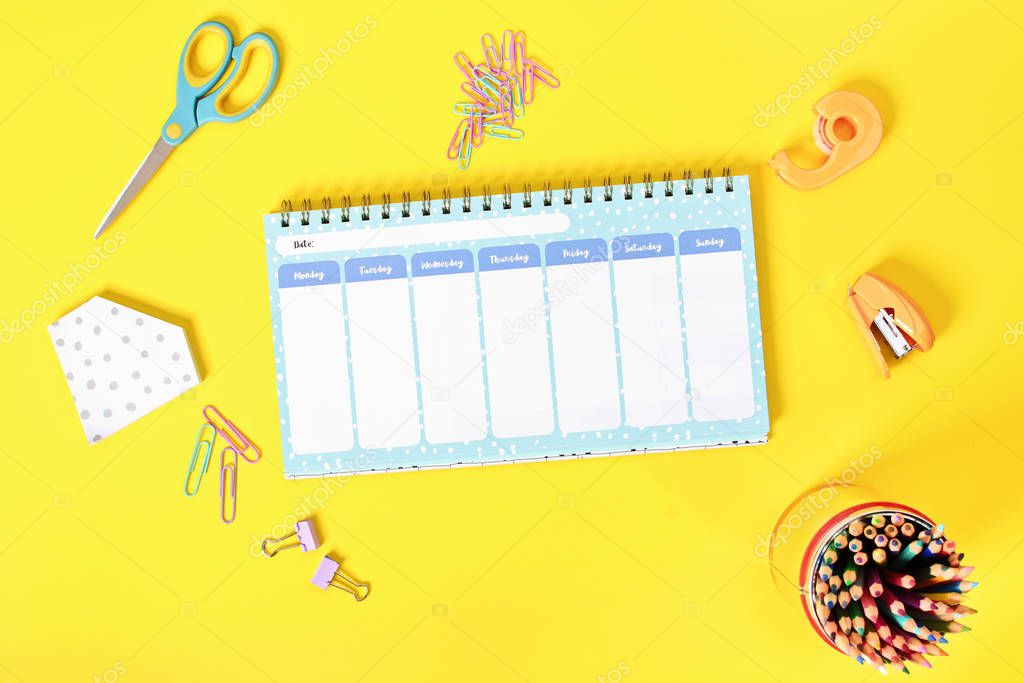 Close up shot of a blank weekly schedule planer with scattered office supplies on yellow background.