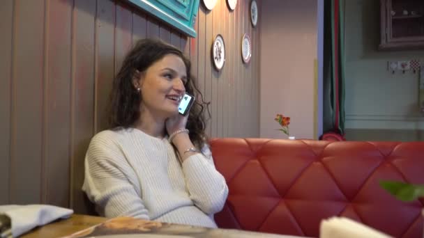 Young woman speaks on the phone and smiling looks at the camera. Rest in a cafe. — Stock Video