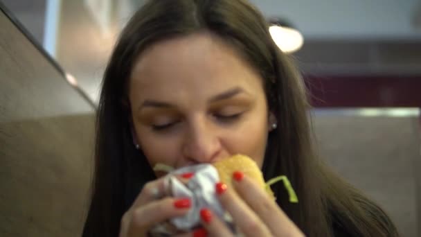 Голодна дівчина їсть гамбургер. Жінка кусає чізбургер у ресторані швидкого харчування — стокове відео