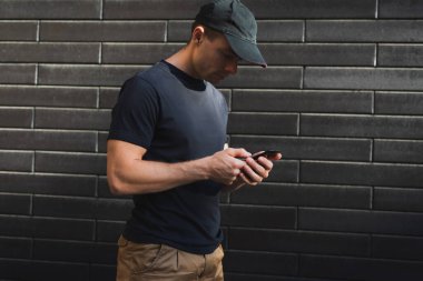 Siyah tişörtlü ve beysbol şapkalı arka planda bir adam akıllı telefona bakar.