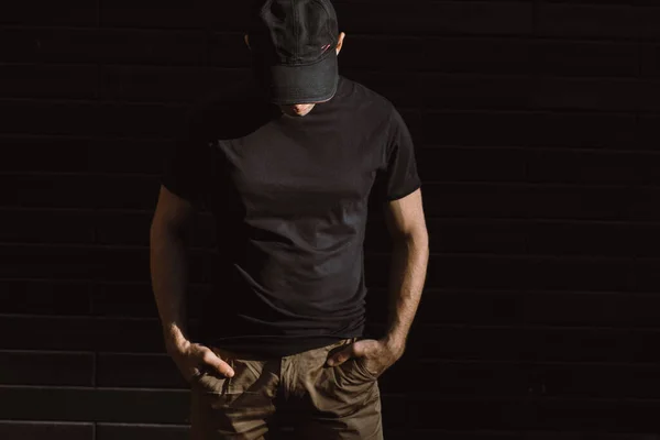 Man in black t-shirt and baseball cap posing against a black wall