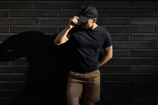 Man in black t-shirt and baseball cap posing against a black wall