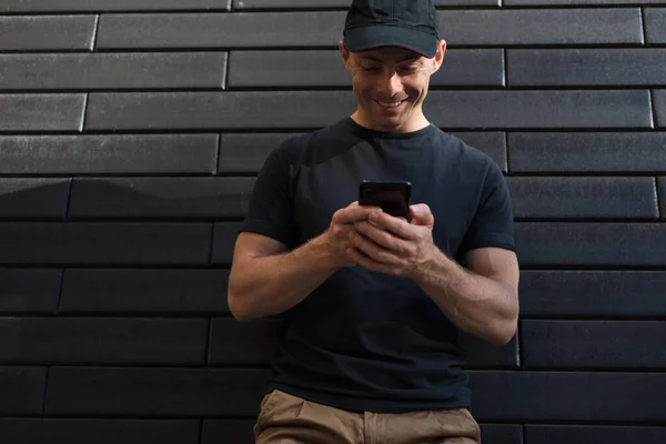 A man on a wall background in a black t-shirt and baseball cap looks into a smartphone