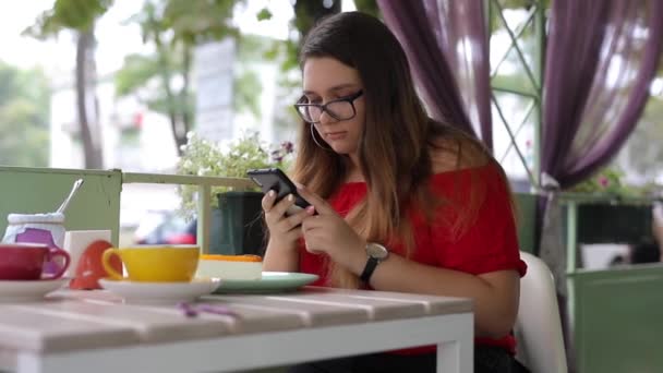 Menina bonito se comunica em um smartphone em uma mesa em um café — Vídeo de Stock