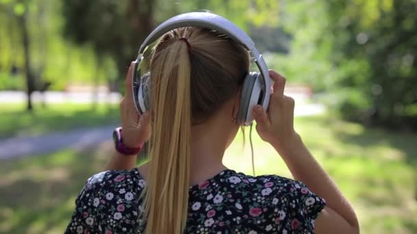 Mujer se pone auriculares y camina hacia adelante en el parque — Vídeos de Stock