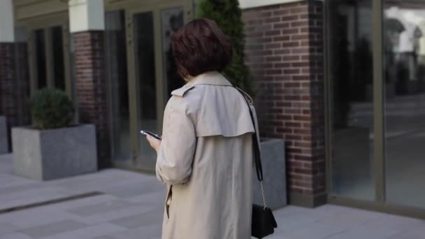 Милая женщина болтает в смартфоне в плаще, вид сзади — стоковое видео